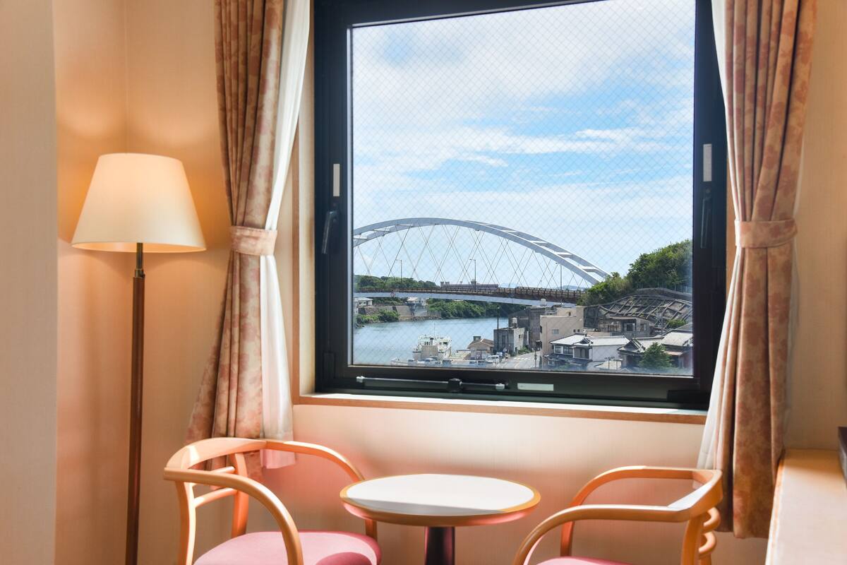 A room with a window view of a bridge, a lamp, and two chairs.