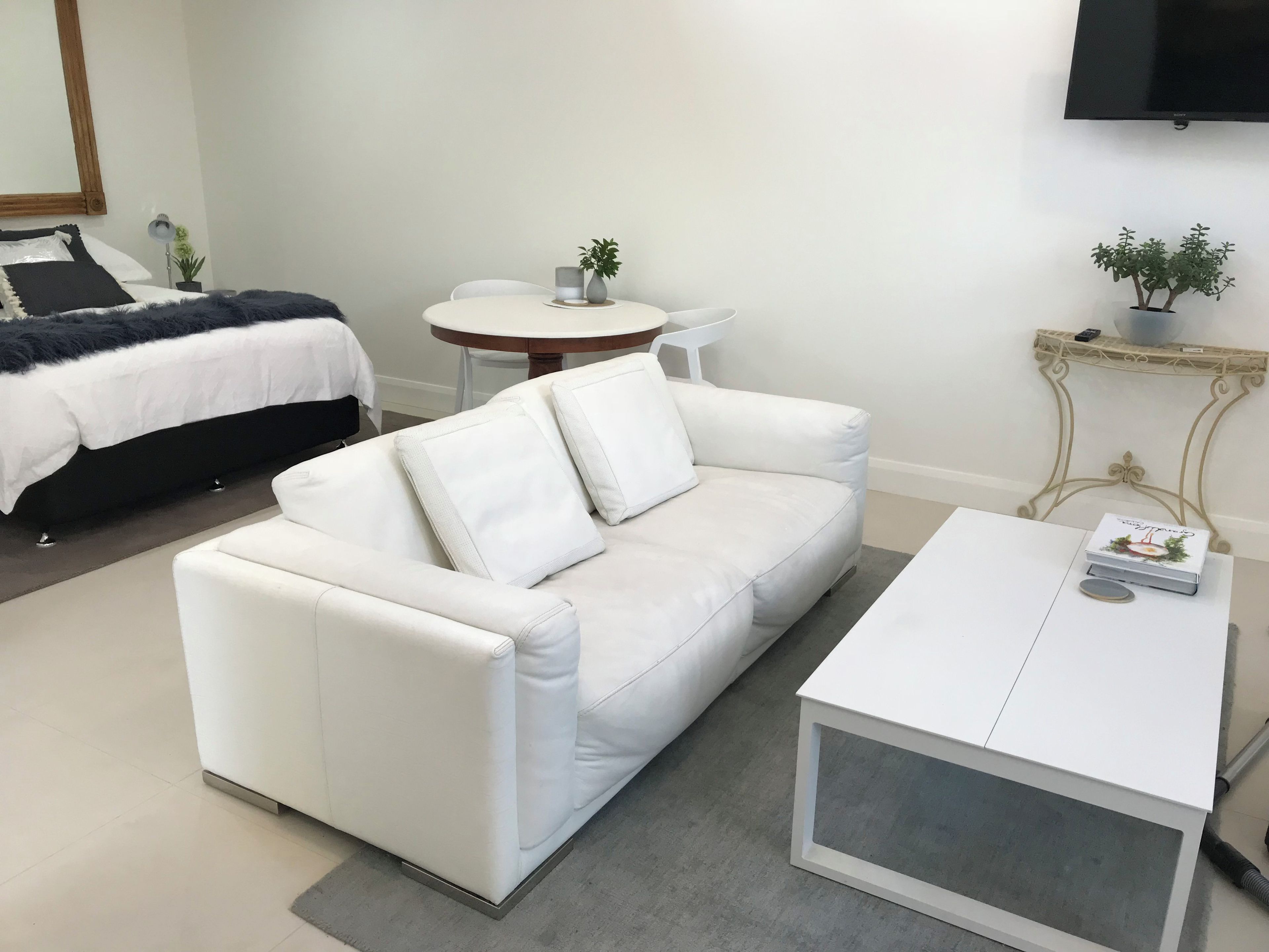 Un soggiorno moderno con un divano bianco, un tavolino, un tavolo rotondo con sedie, un letto con copripiumino bianco e nero, e una TV montata a parete.