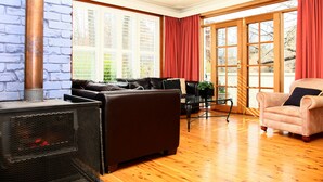 Ein Wohnzimmer mit Kamin, einer Couch, einem Sessel und großen Fenstern mit roten Vorhängen.