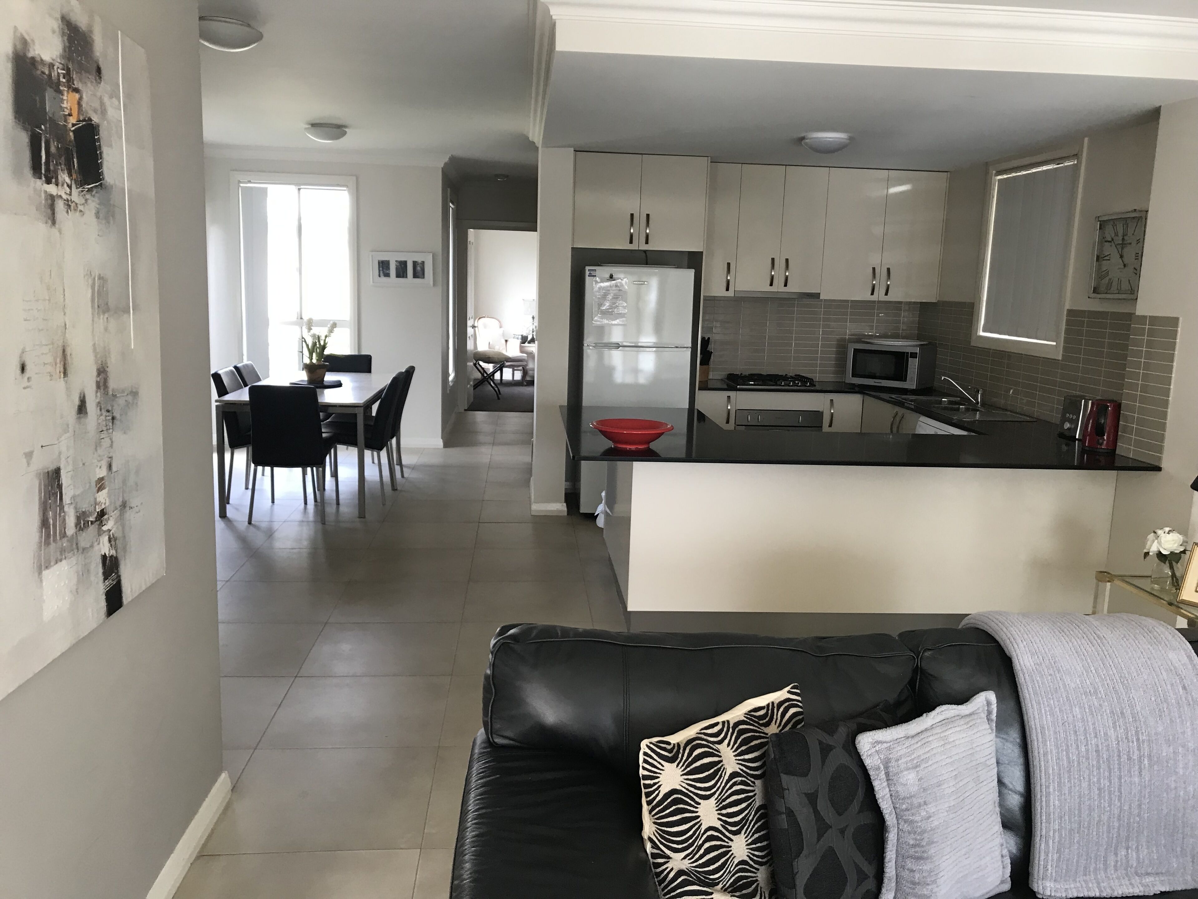 Una cocina moderna con armarios blancos, encimera negra y un tazón rojo. Hay un comedor con mesa y sillas, y una sala de estar con sofá negro y cojines estampados.