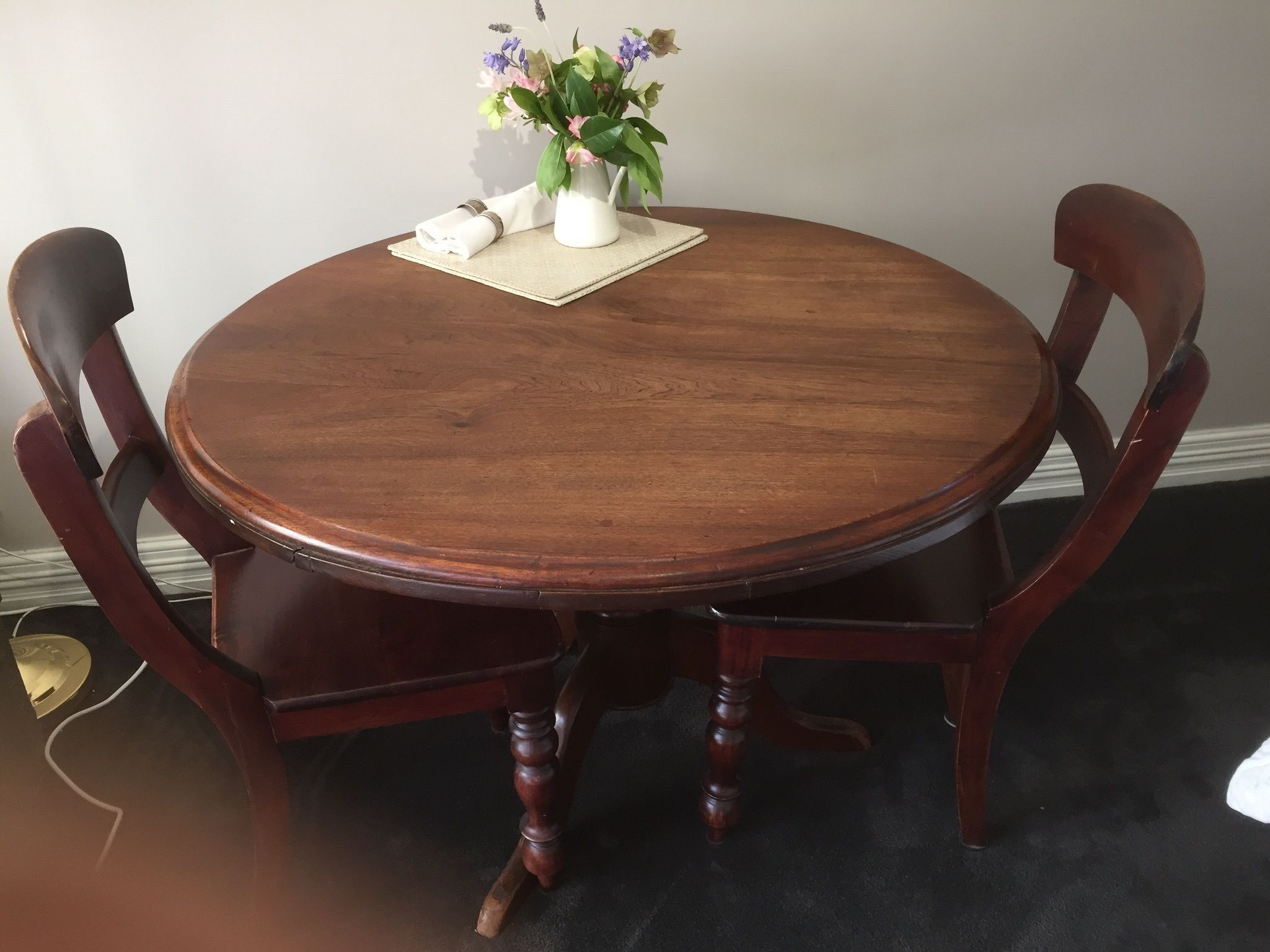 A round wooden dining table with four chairs, a vase with flowers, and a placemat on the table.