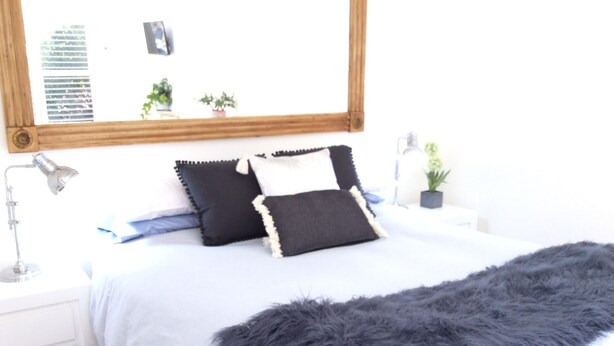 Ein ordentlich bezogenes Bett mit einer flauschigen, grauen Decke, einer Nachttischlampe und einem kleinen Topfpflanz auf einem weißen Nachttisch.
