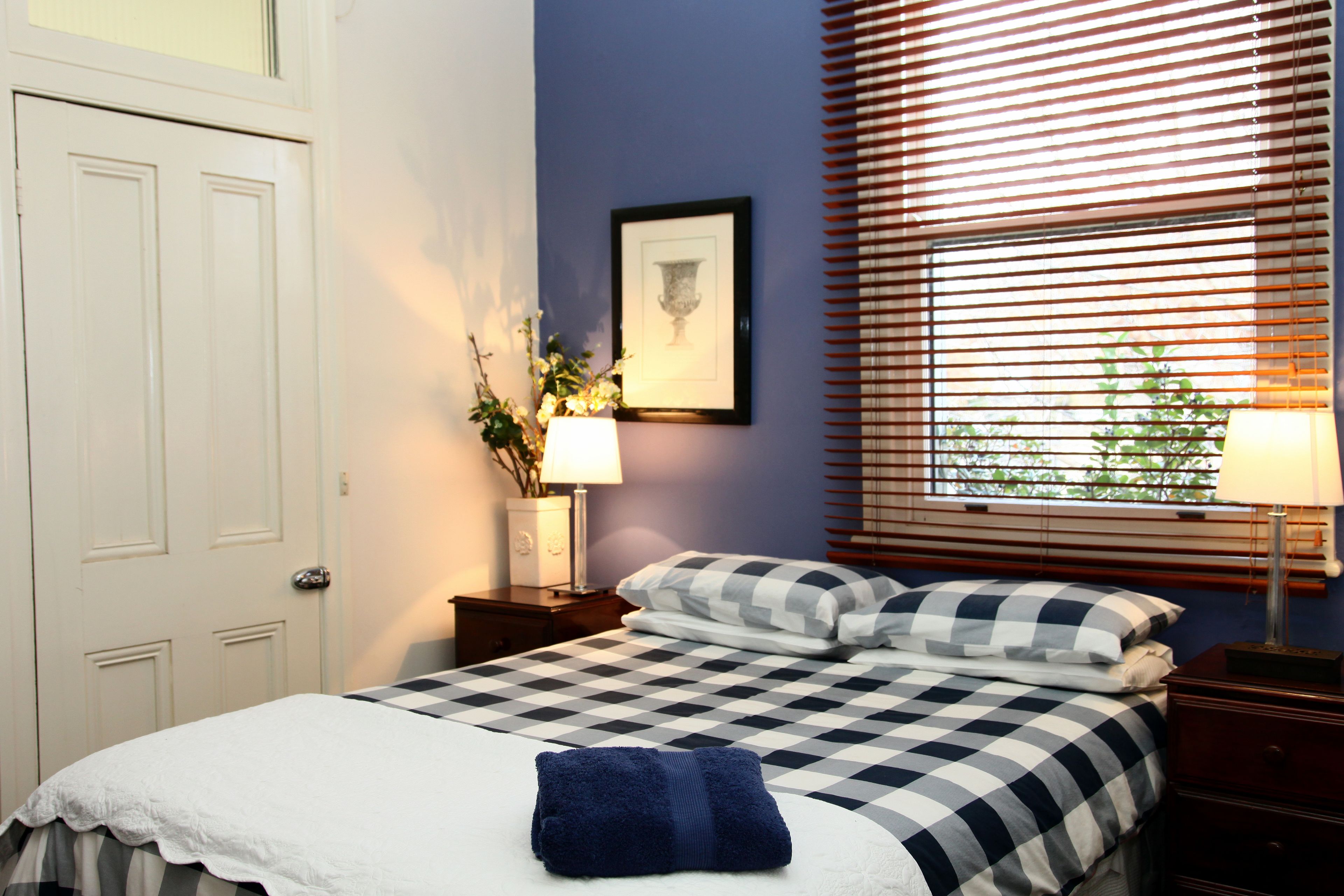 A neatly made bed with a checkered bedspread, a blue towel, and a framed picture on the wall.