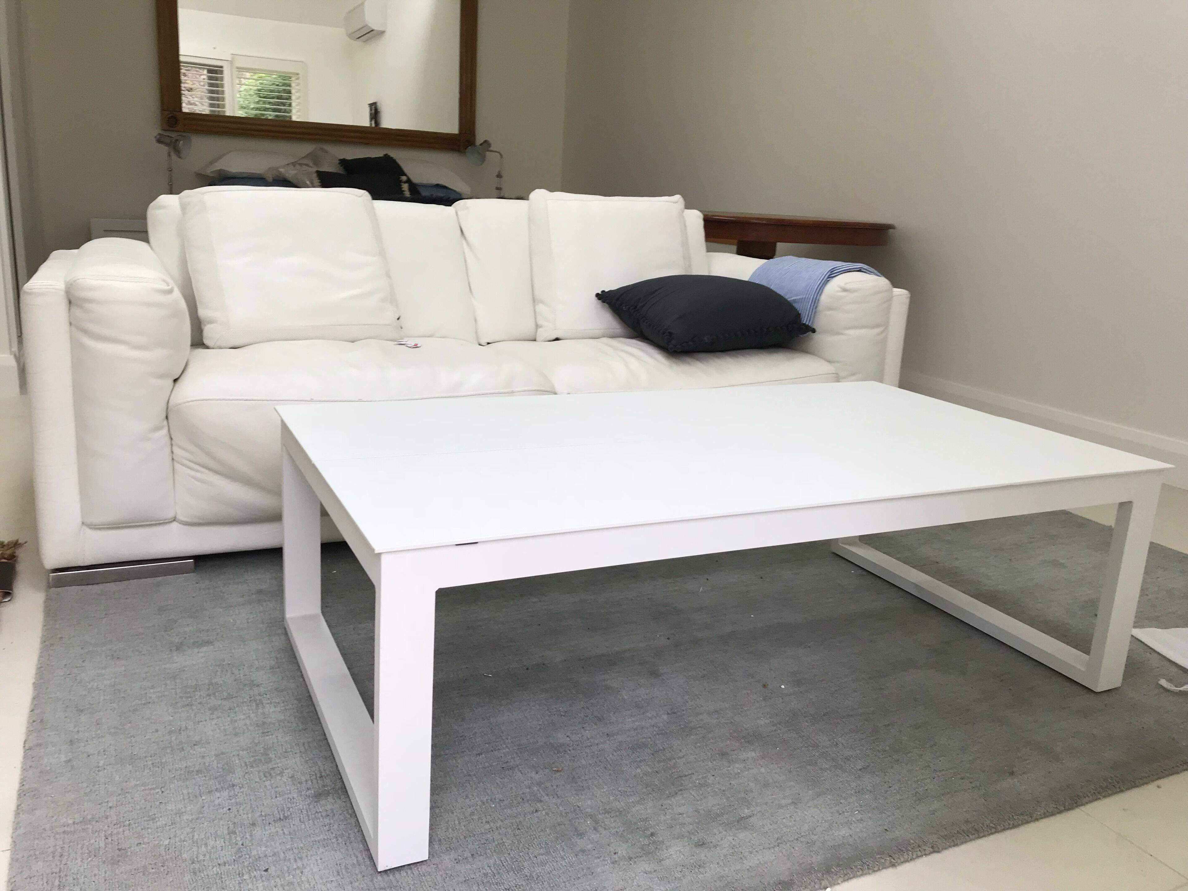 A white leather sofa with a black pillow, a white coffee table, and a grey rug in a room with a mirror on the wall.