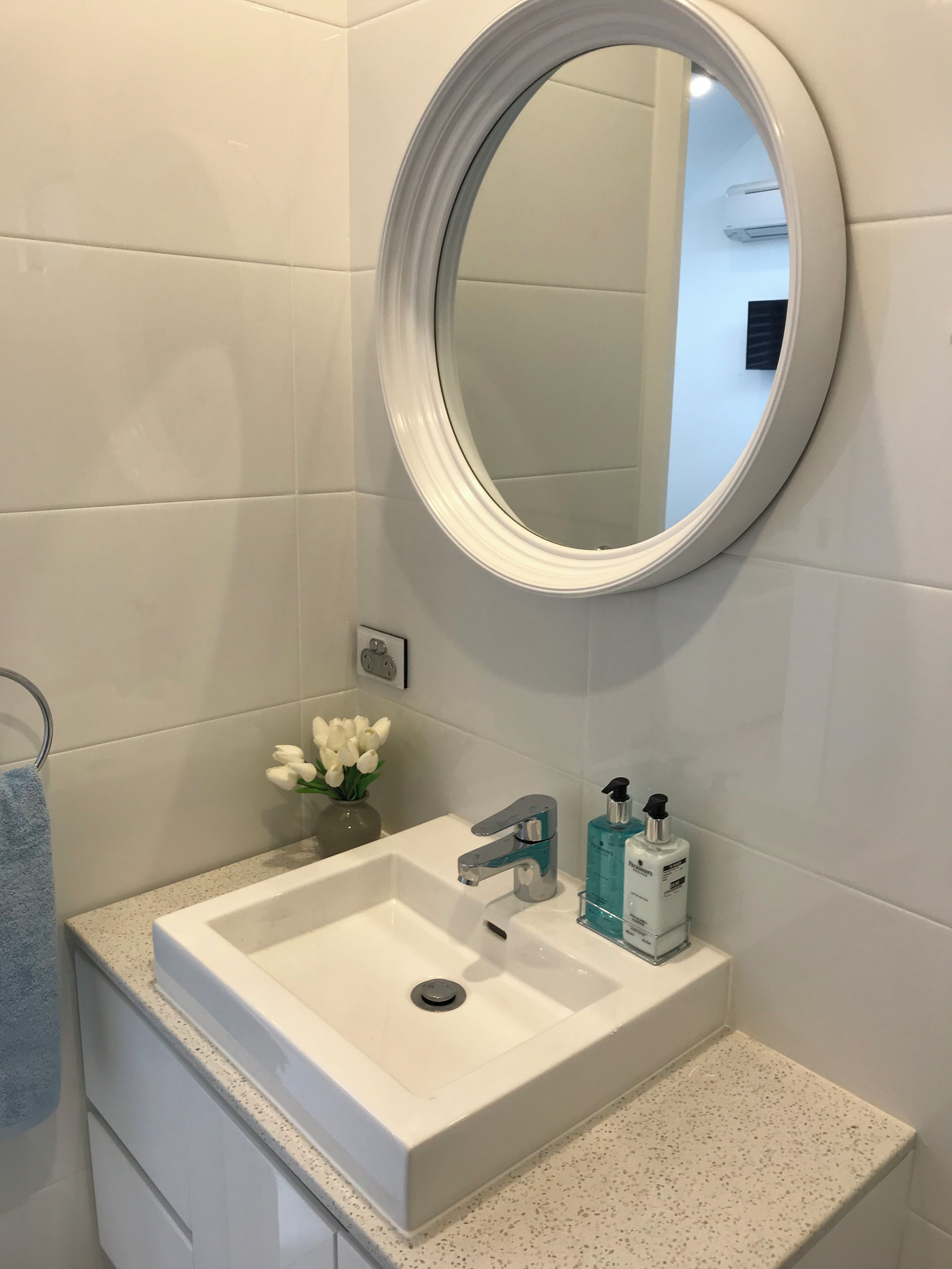 A bathroom with a white sink, a round mirror, a towel rack, and a vase with white flowers.
