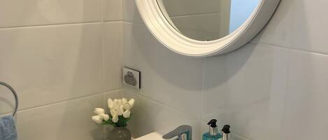 A bathroom with a white sink, a round mirror, a towel rack, and a vase with white flowers.
