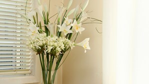 Ein Zimmer mit einem beige gefärbten Sofa, einem gemusterten Kissen, einem kleinen Tisch mit Blumen in einer Vase und einem Fenster mit Jalousien.
