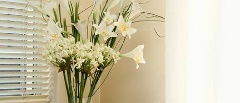 Una habitación con un sofá color beige, un cojín estampado, una mesita con un jarrón de flores y una ventana con persianas.