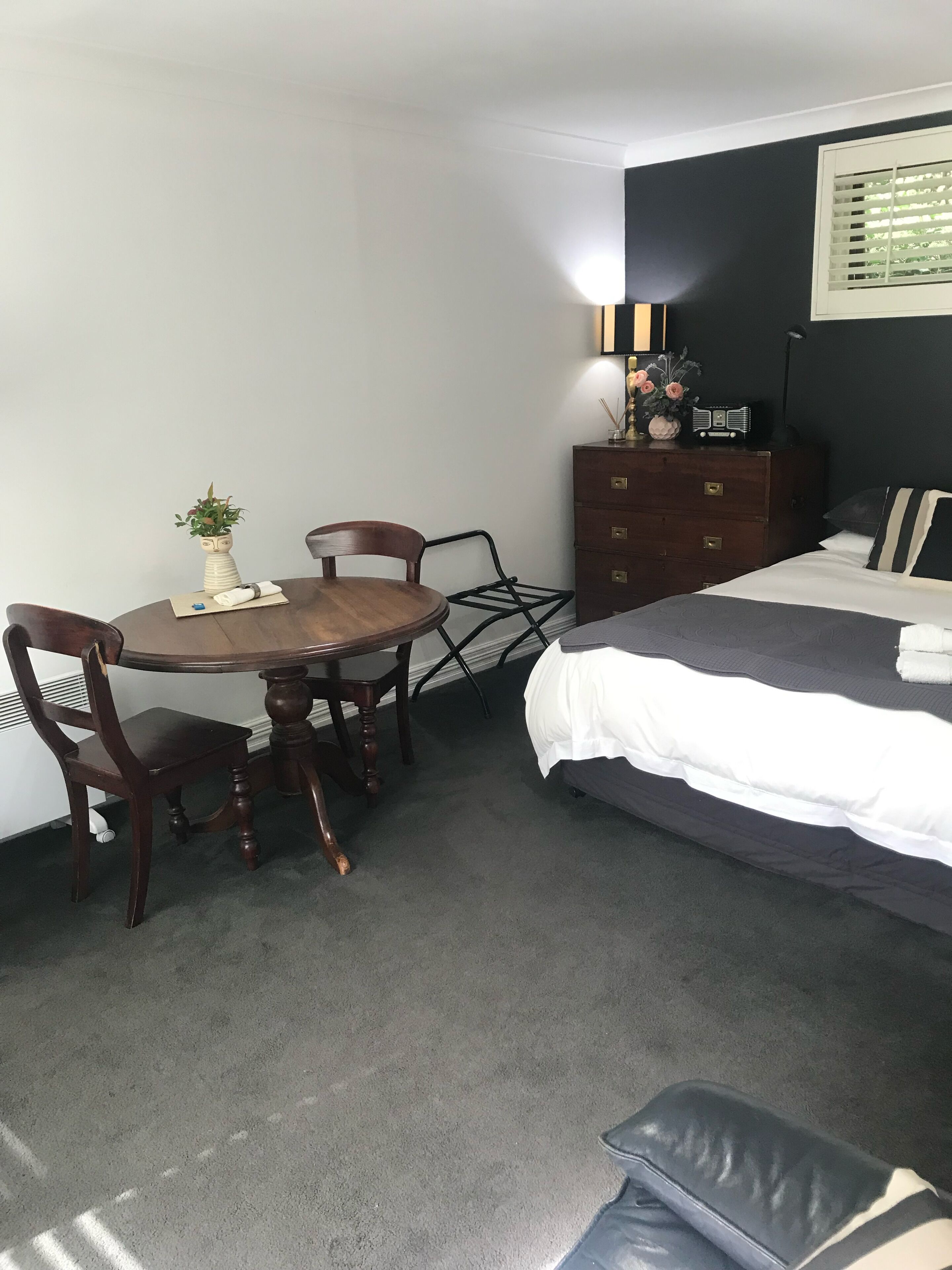 Une chambre d’hôtel compacte comprenant une table de salle à manger ronde en bois, deux chaises, un lit simple avec une couette grise et blanche, une commode avec une radio et une petite plante en pot.