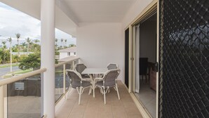 Un balcon avec une table et des chaises blanches, une balustrade en verre et une vue sur des palmiers et des maisons.