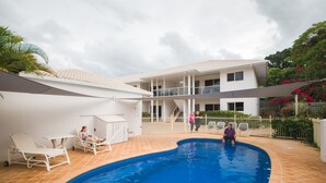 Une piscine dotée de chaises longues et d’un espace de repos couvert, entourée d’un bâtiment blanc avec un toit en tuiles et un jardin clôturé.