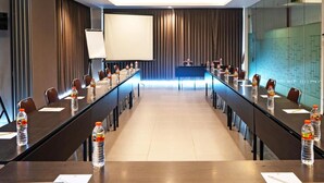 Een congreszaal met een lange tafel, stoelen, waterflesjes, een scherm voor een beamer en een glazen scheidingswand.