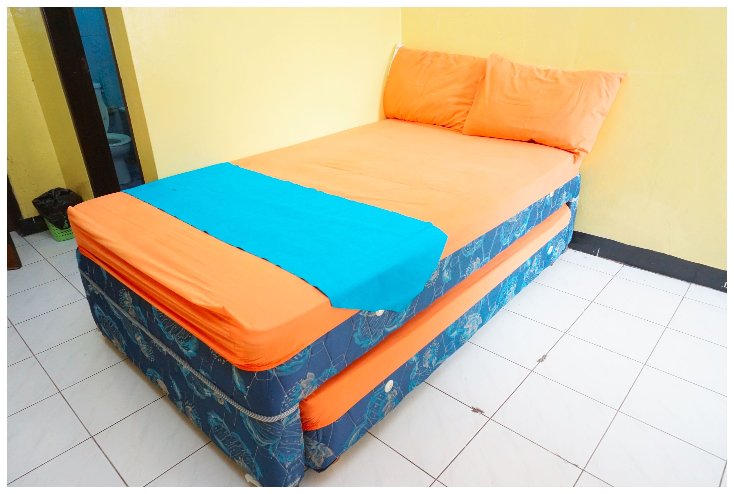 Un lit avec une couvre-lit orange et bleue, une poubelle verte et un mur jaune.