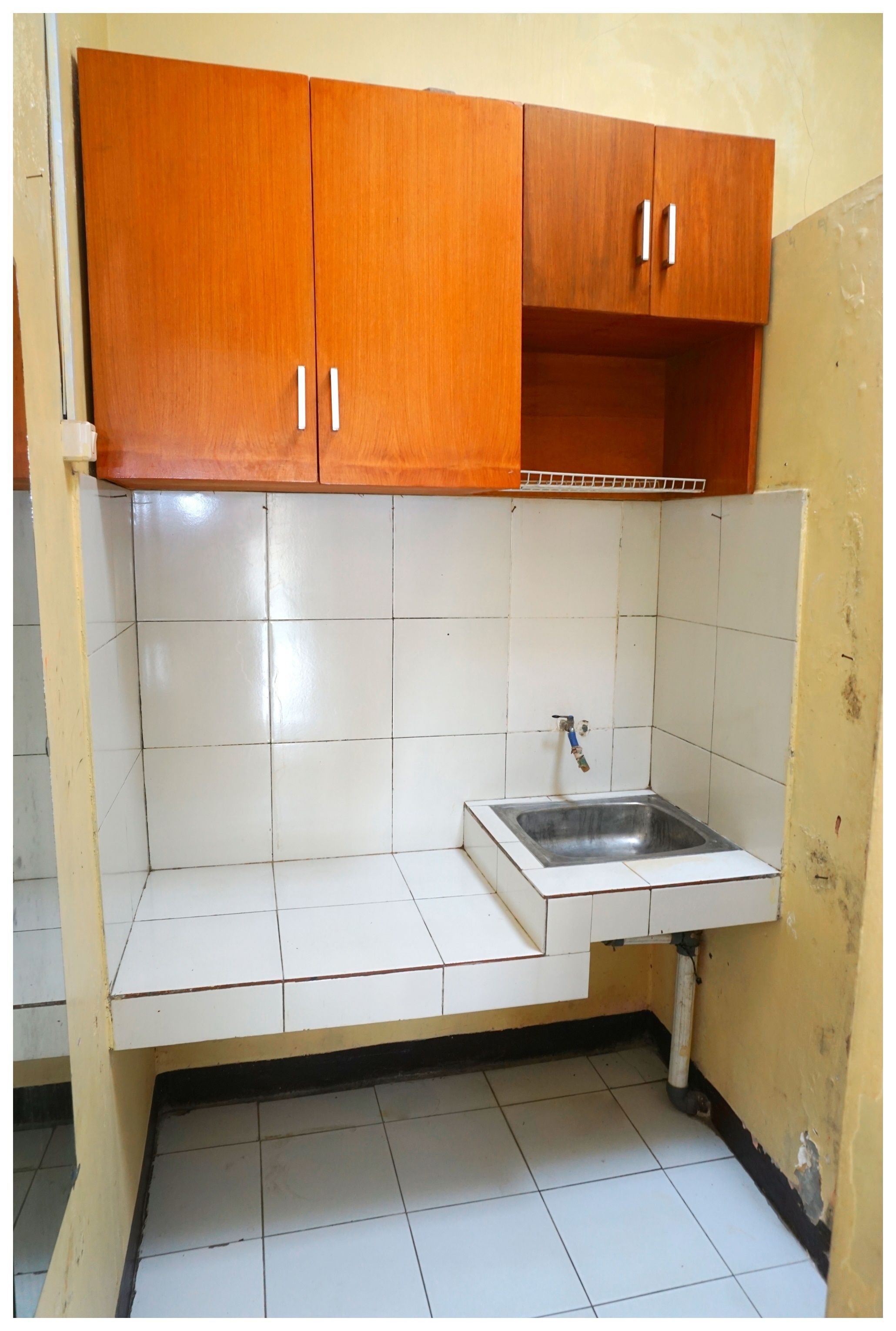 A kitchen with white tiled walls, a stainless steel sink, and wooden cabinets.
