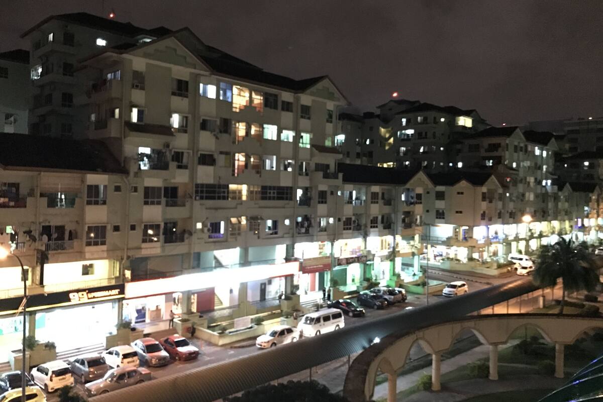 Un edificio de varios pisos con ventanas iluminadas por la noche, un estacionamiento con varios coches y un puente peatonal en primer plano.