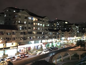 Un immeuble à plusieurs étages dont les fenêtres sont illuminées la nuit, un parking avec plusieurs voitures et un pont piétonnier au premier plan.