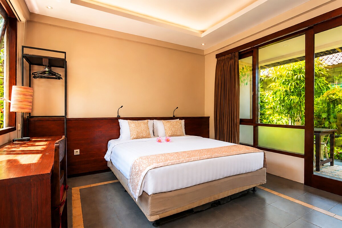 Habitación de hotel con cama, mesita de noche, lámpara y vistas a zonas verdes a través de amplios ventanales.