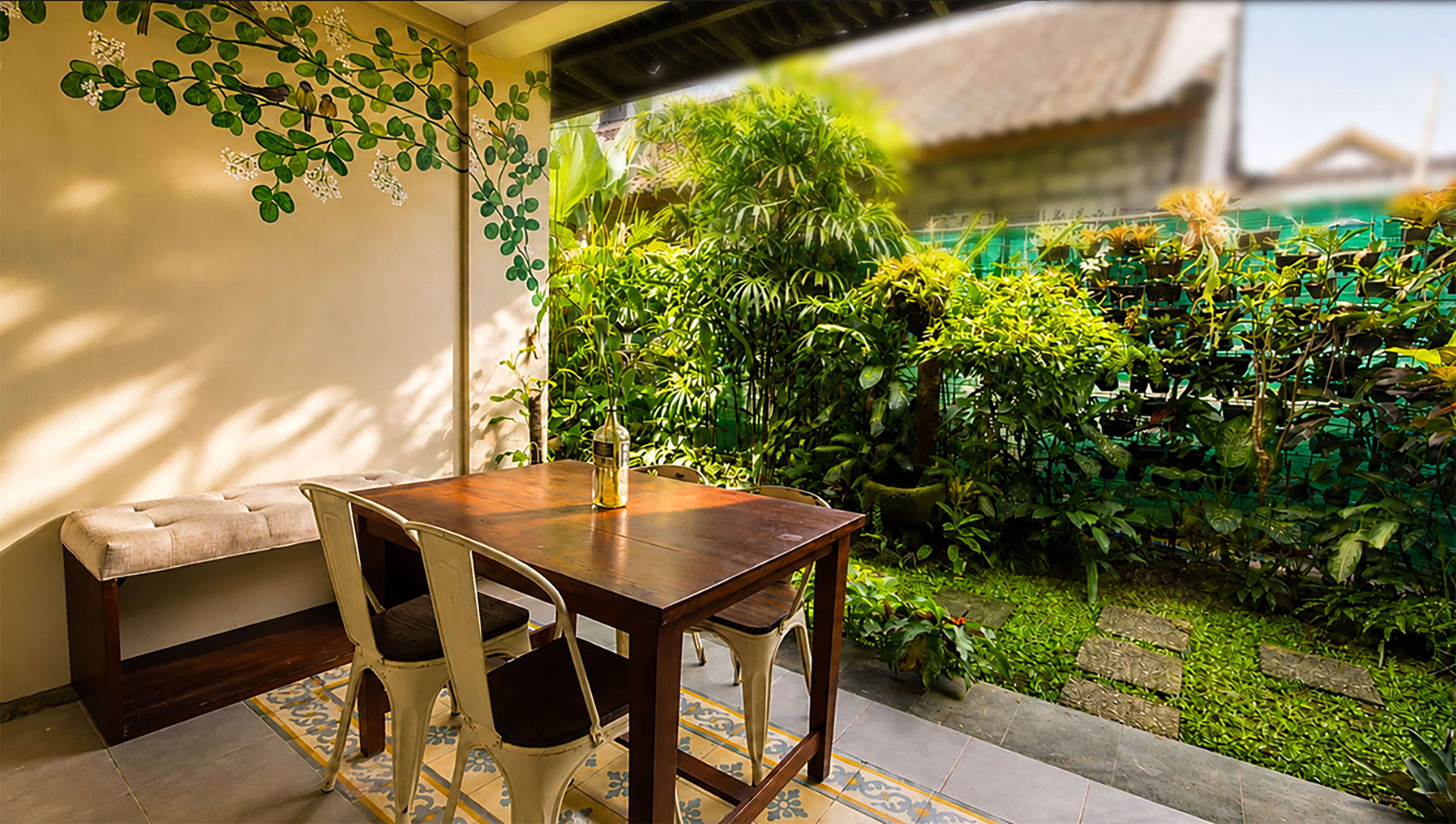 Un espace repas comprenant une table et des chaises en bois, un banc, et donnant sur un jardin luxuriant avec différentes plantes et une clôture verte.