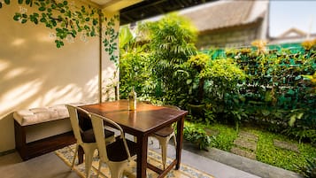 Un espace repas comprenant une table et des chaises en bois, un banc, et donnant sur un jardin luxuriant avec différentes plantes et une clôture verte.