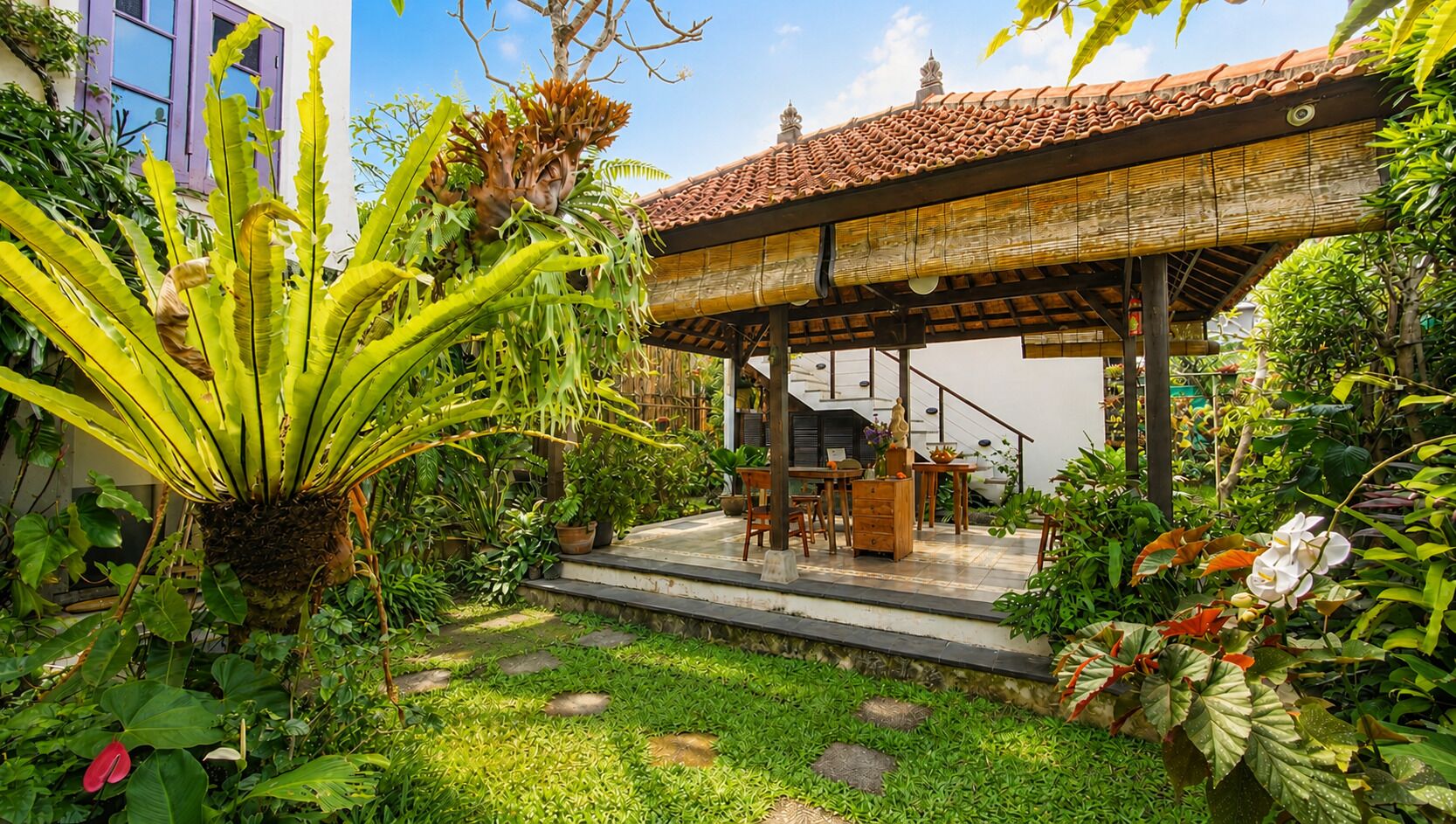 A covered outdoor seating area with a staircase leading to a building, surrounded by lush greenery and plants.