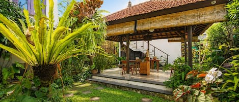 Un espacio de estar techado al aire libre con una escalera que lleva a un edificio, rodeado de exuberante vegetación y plantas.