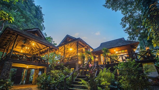 A rustic lodge with multiple wooden structures connected by bridges, surrounded by dense vegetation.