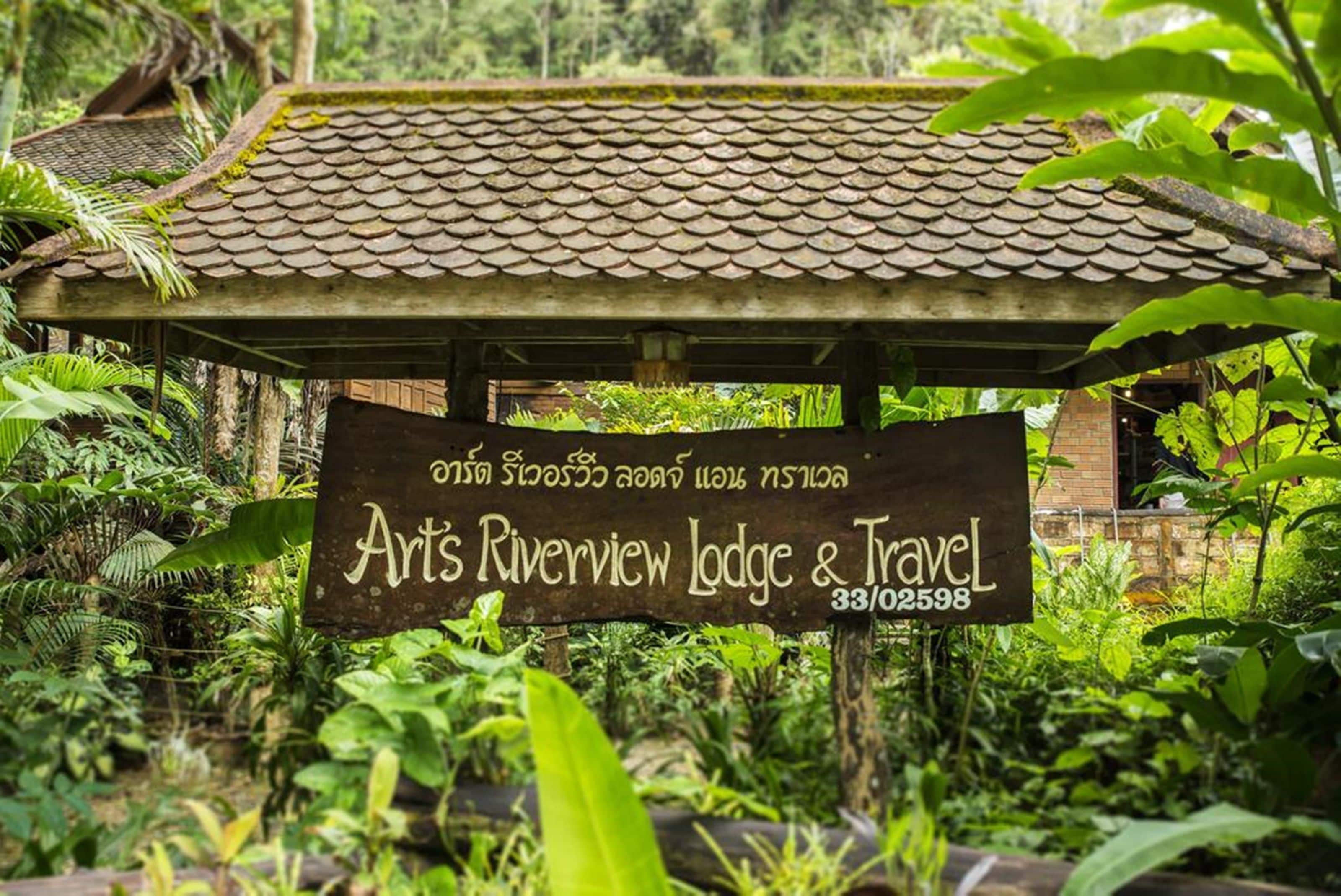 Skilt for Arts Riverview Lodge & Travel med et traditionelt tag og omgivet af frodig vegetation.