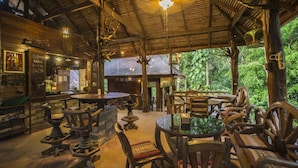 A rustic wooden interior with round tables and chairs, a bar area, and a view of lush greenery outside.