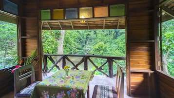 Una cabaña de madera con balcón que da a un bosque. Se ve una mesa con mantel floral y dos sillas.