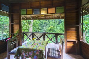 Una cabaña de madera con balcón que da a un bosque. Se ve una mesa con mantel floral y dos sillas.