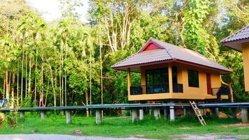 A single-story house with a red roof, supported by concrete stilts, surrounded by lush greenery and tall trees.