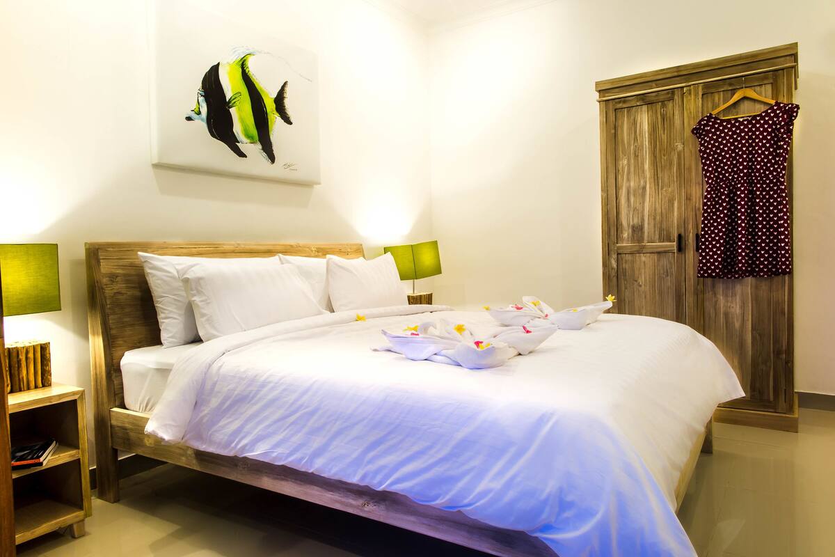 A neatly made bed with white linens and a floral arrangement, a wooden headboard, a wooden wardrobe, and a green wall lamp.