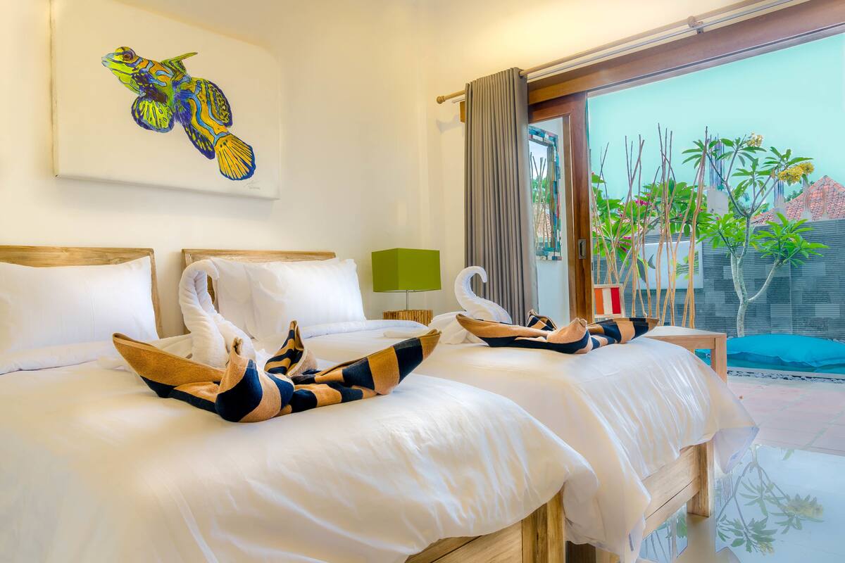 A hotel room with two beds, a painting on the wall, a window with a view of the outside, and a small table with a lamp.