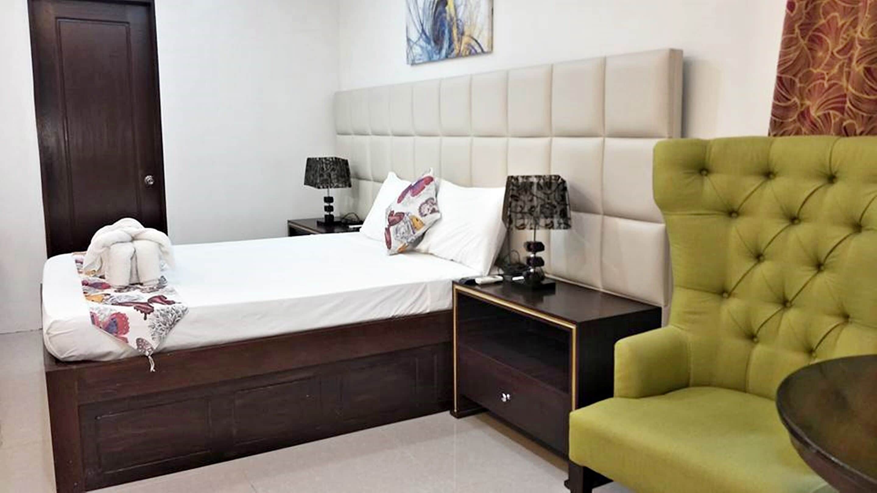 A hotel room with a bed, two bedside tables, a green armchair, and a painting on the wall.