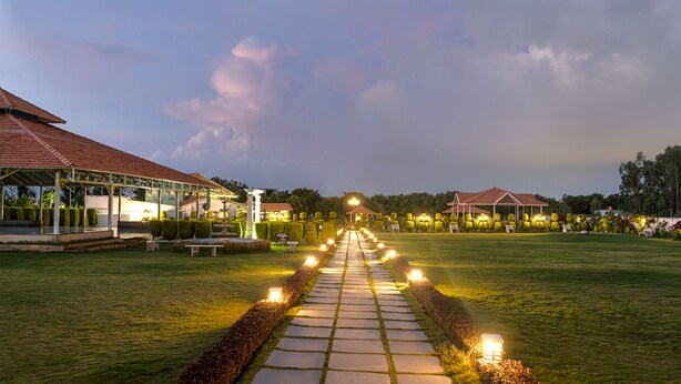A pathway leading to a building with a red roof, surrounded by well-maintained hedges and illuminated by ground lights. - Fiestaa Resort n Events Venue (Bengaluru)