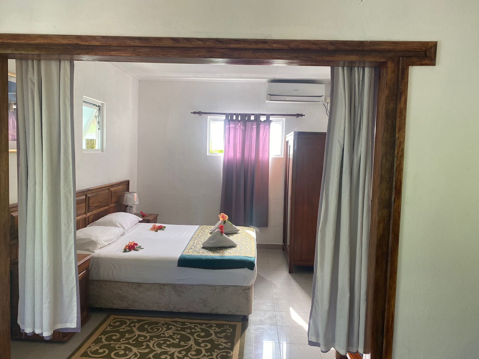 A hotel room with a bed, a nightstand, an air conditioning unit, and a window with curtains.