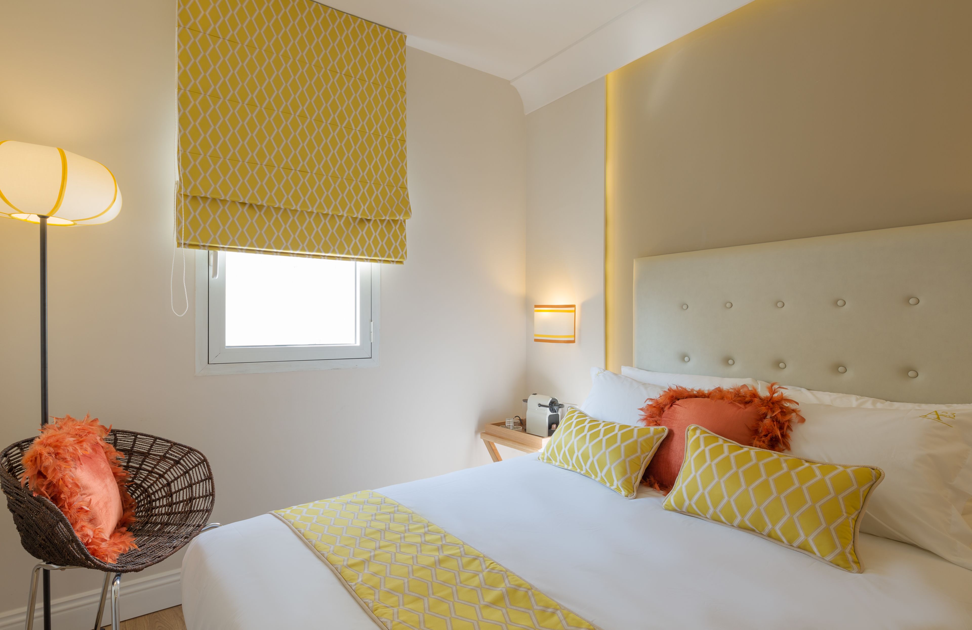 A hotel room with a bed, a chair, a bedside table, a lamp, and a window with a patterned blind.