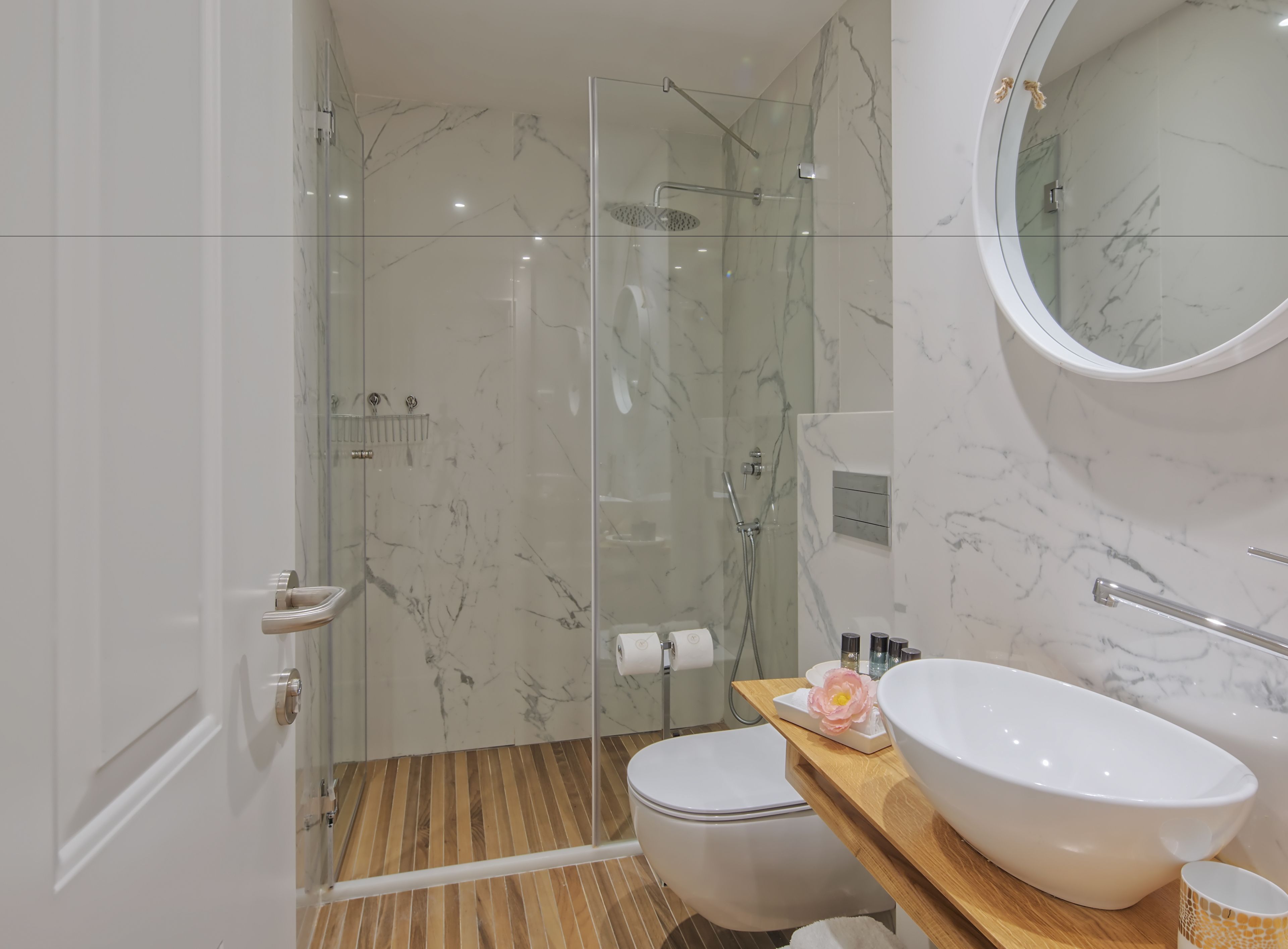 A modern bathroom with a glass-enclosed shower, a white toilet, and a wooden vanity with a large white basin and a small decorative flower.