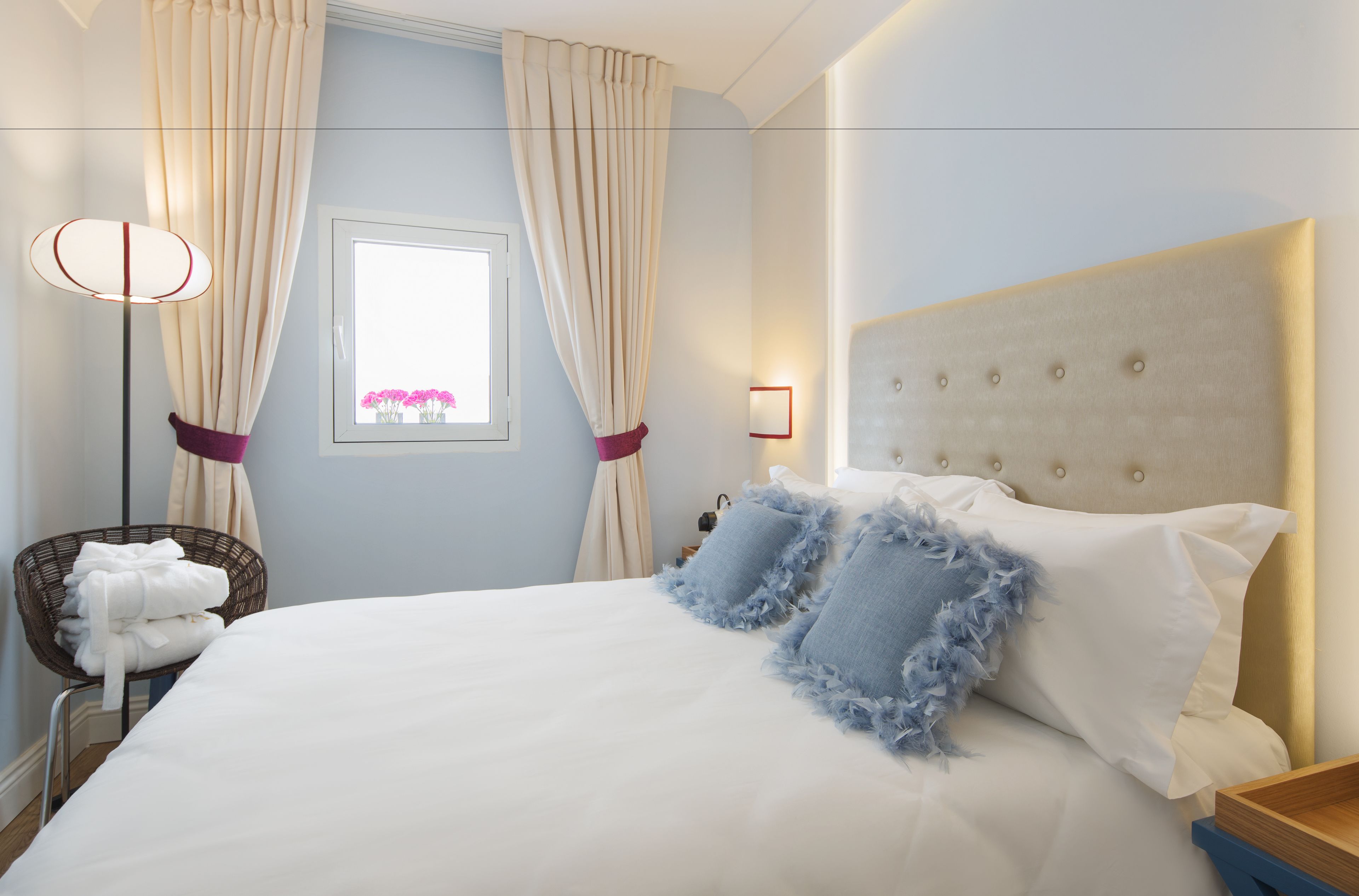Ein ordentlich bezogenes Bett mit weißen Laken und blauen Kissen, ein Nachttisch mit Lampe sowie ein Fenster mit Vorhängen.