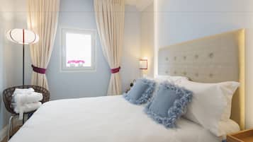 Ein ordentlich bezogenes Bett mit weißen Laken und blauen Kissen, ein Nachttisch mit Lampe sowie ein Fenster mit Vorhängen.