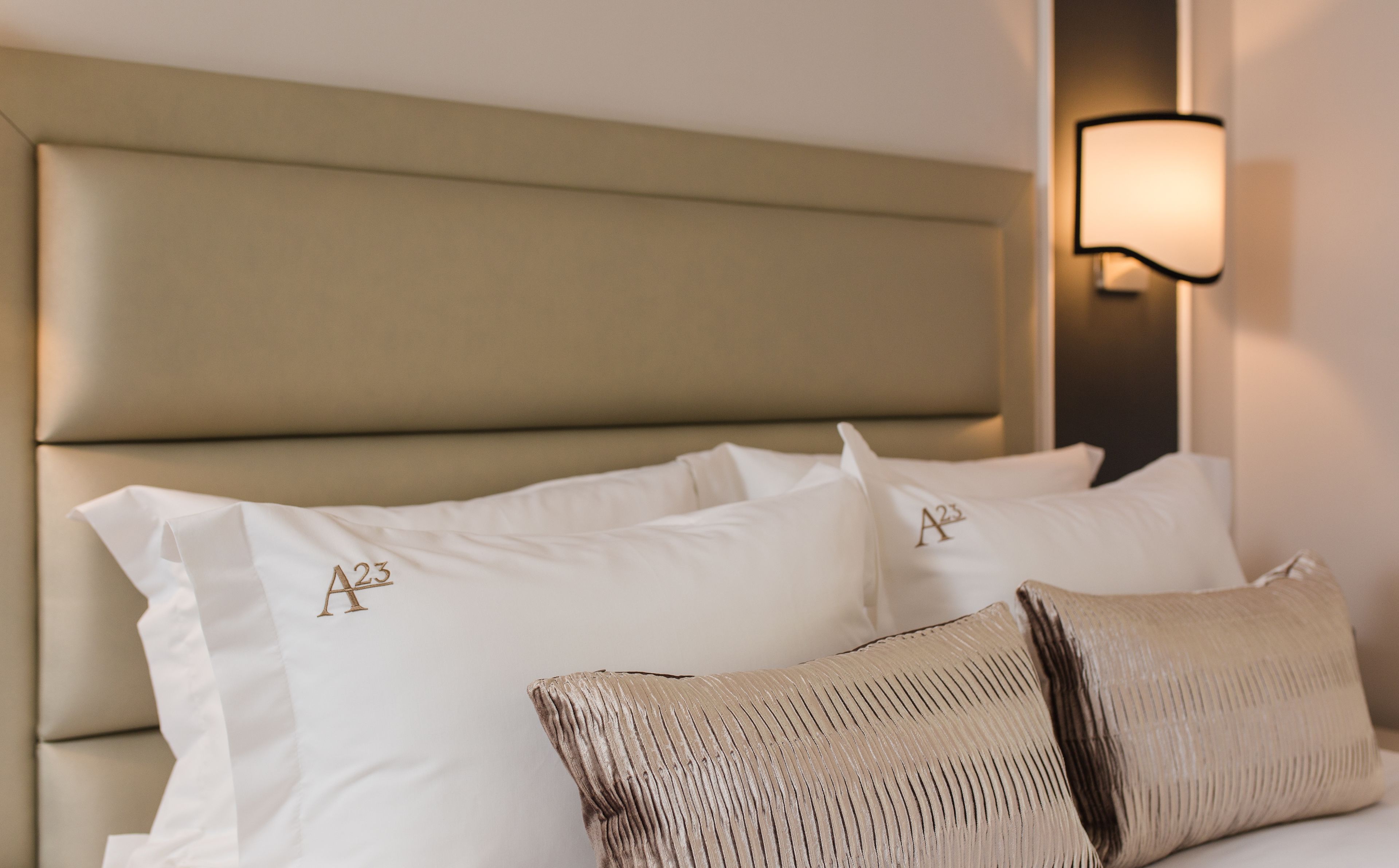 A bed with a headboard, two pillows with embroidered initials, a textured throw pillow, and a wall-mounted light fixture.