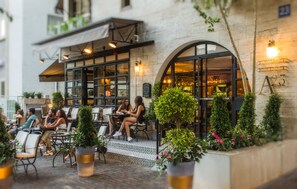 Un espace extérieur aménagé avec des tables et des chaises, des plantes en pot et un bâtiment sur lequel est apposée une enseigne indiquant « A23 ».