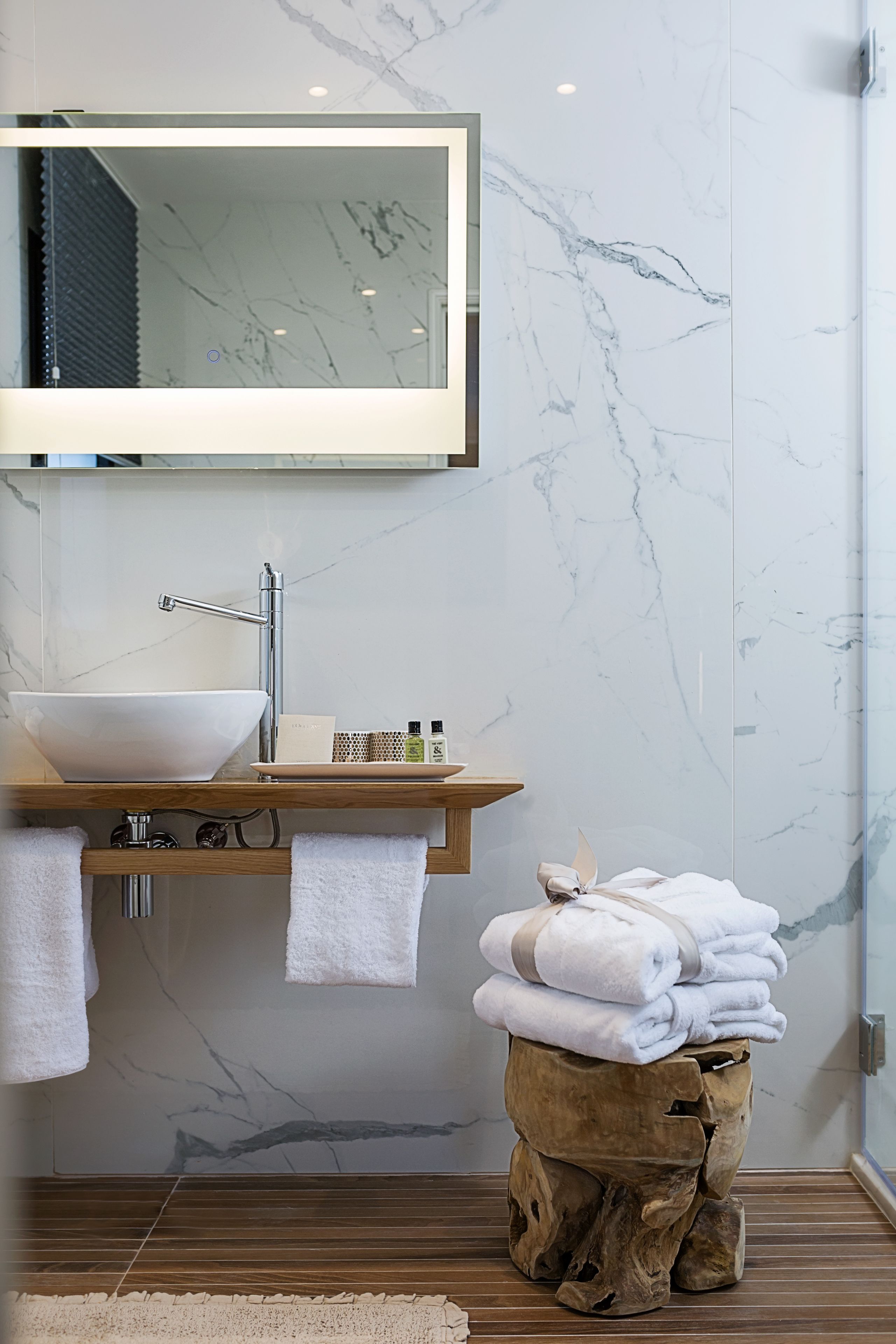 Une salle de bain avec un lavabo en marbre, un meuble-lavabo en bois, un miroir et une pile de serviettes posées sur un tabouret en bois.