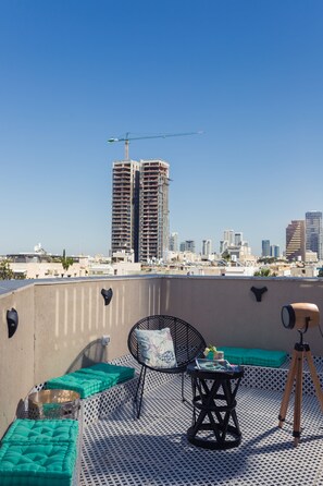 Une terrasse sur le toit offrant une vue sur les toits de la ville, notamment sur un grand immeuble en construction. On y trouve des espaces de détente en plein air avec des coussins et une table.
