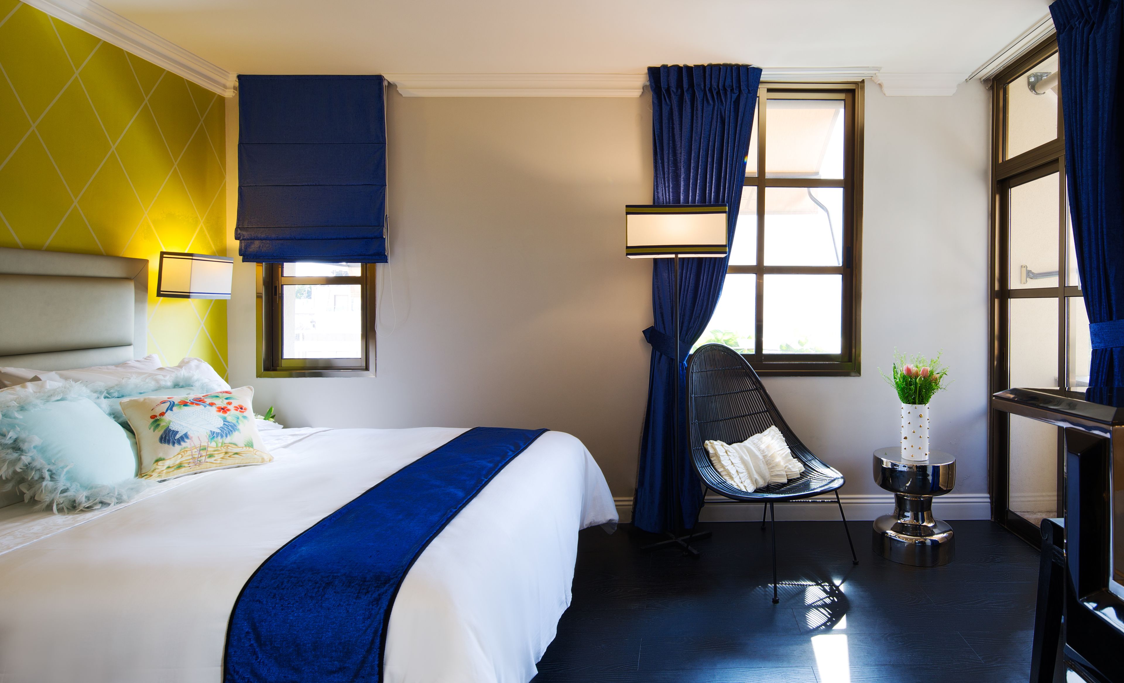 A hotel room with a bed, a chair, a small table with a vase, and a window with curtains.
