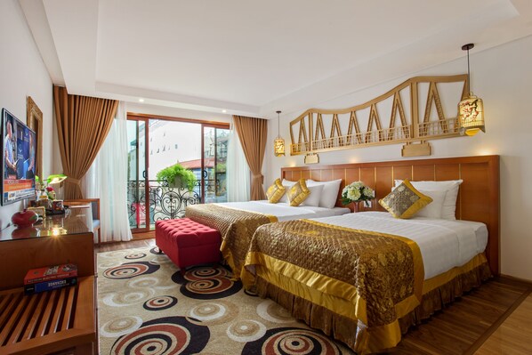 Two double beds with gold and white bedding, a red ottoman, and a desk with a lamp and television.