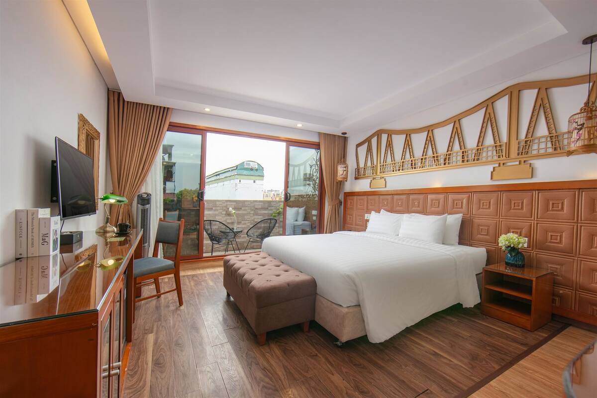 A hotel room with a bed, a desk with a TV, a chair, a small table, and a balcony with a view.
