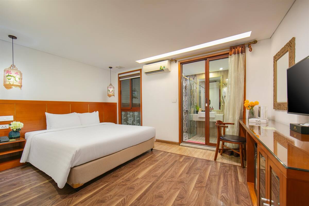 A hotel room with a bed, wooden flooring, a desk with a chair, a television, and a sliding glass door leading to a bathroom.