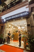 Exterior del Hotel Golden Holiday, con fachada de mármol, alfombra roja en la entrada y dos empleados de pie allí.