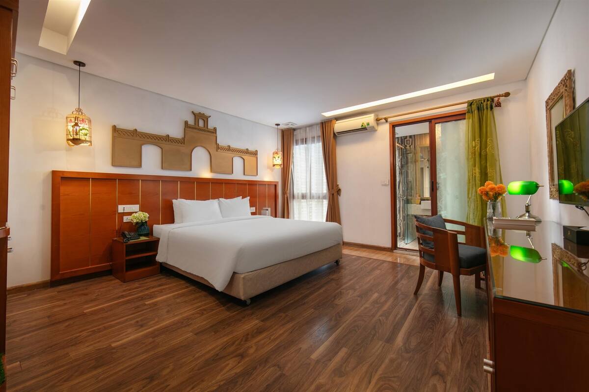 A hotel room with a wooden floor, a bed with white linens, a desk with a chair, and a large mirror.